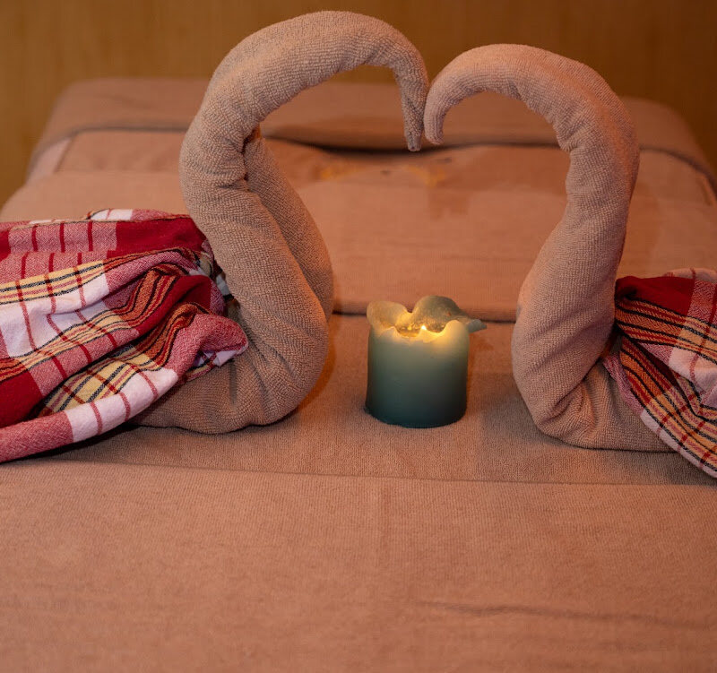 Two towels folded into a heart on a treatment bed, with a lit candle between them.
