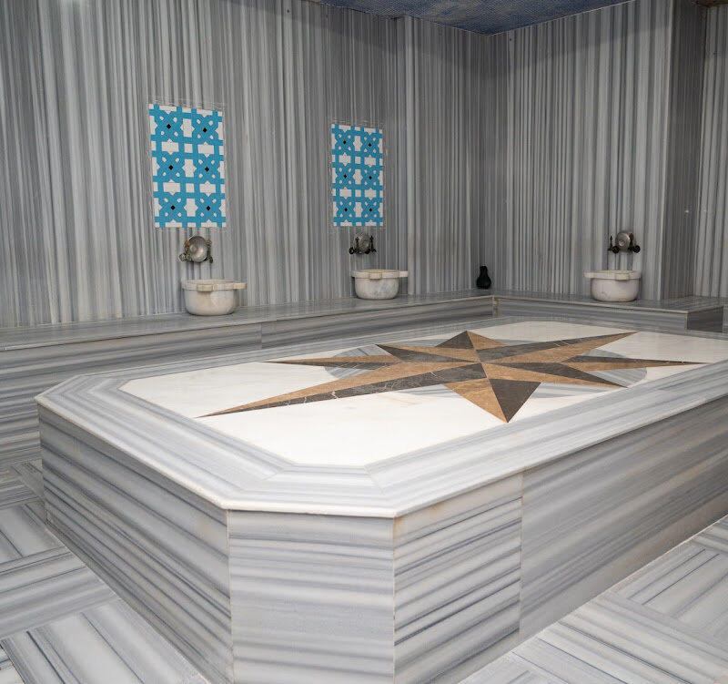 Marble Turkish hamam with a star-inlaid heated slab in the middle and kurna basins under Iznik tile panels.